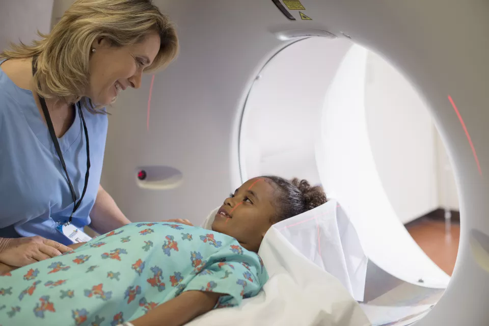 A woman with light toned skin wearing blue scrubs comforts a medium-dark skin toned girl laying on the bed of an MRI machine.