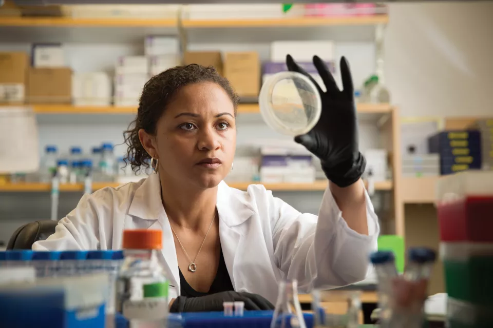 A woman with medium-dark skin tone wearing a lab coat holds a dish up to look at a research sample.