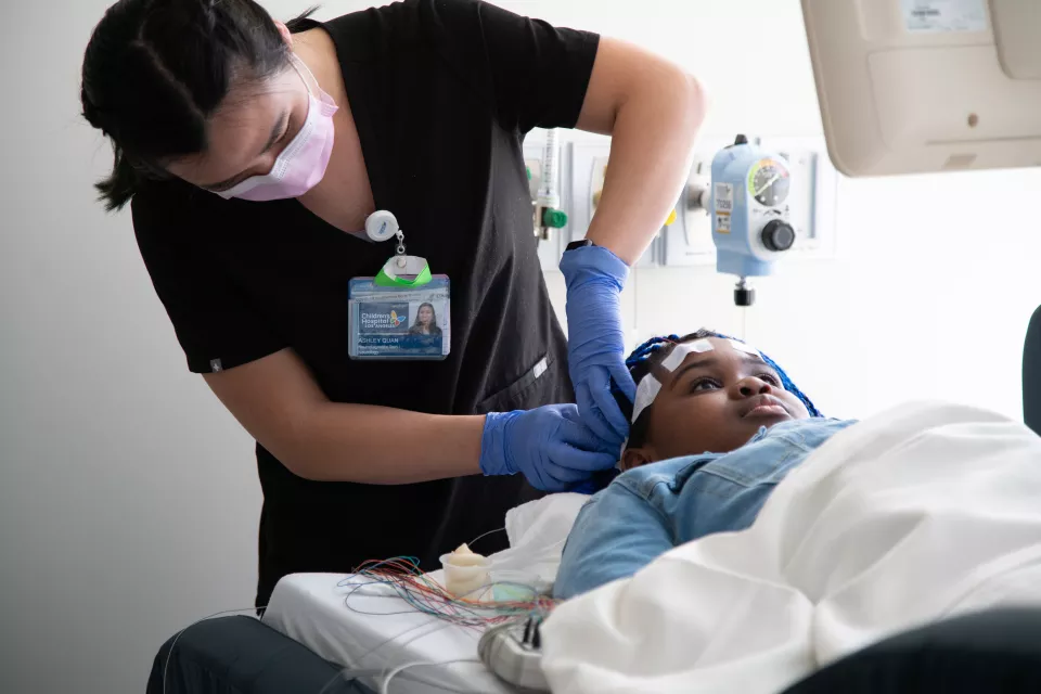 A medium-light skin toned woman in black scrubs and a surgical mask tapes wires to the head of a dark skin toned girl lying on a hospital bed.