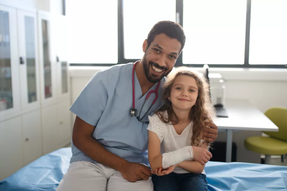 A medium-dark skin toned man in scrubs with a stethscope poses next to a light skin toned girl holding her bandaged arm. Both smile.