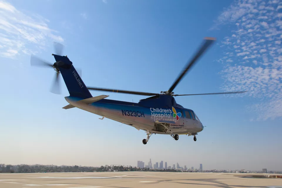 A Sikorsky S-76 helicopter with the Children's Hospital Los Angeles logo hovers above a helipad. The sky is blue and streaked with clouds and downtown L.A. is in the background.