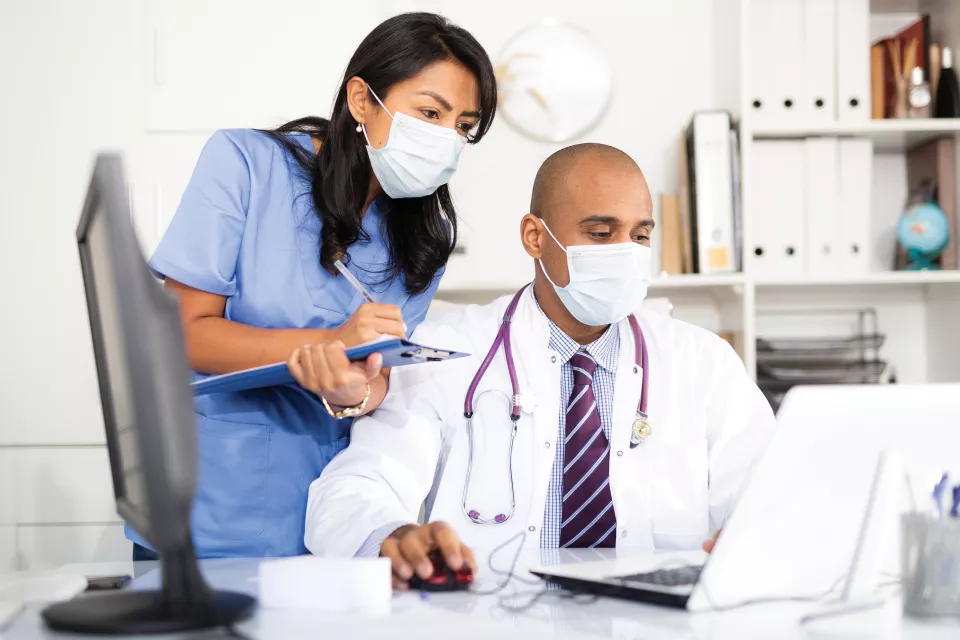 A medium-dark skin toned man in a lab coat uses a laptop. A medium-light skin toned woman in blue scrubs looks over his shoulder, taking notes. Both wear surgical masks. 