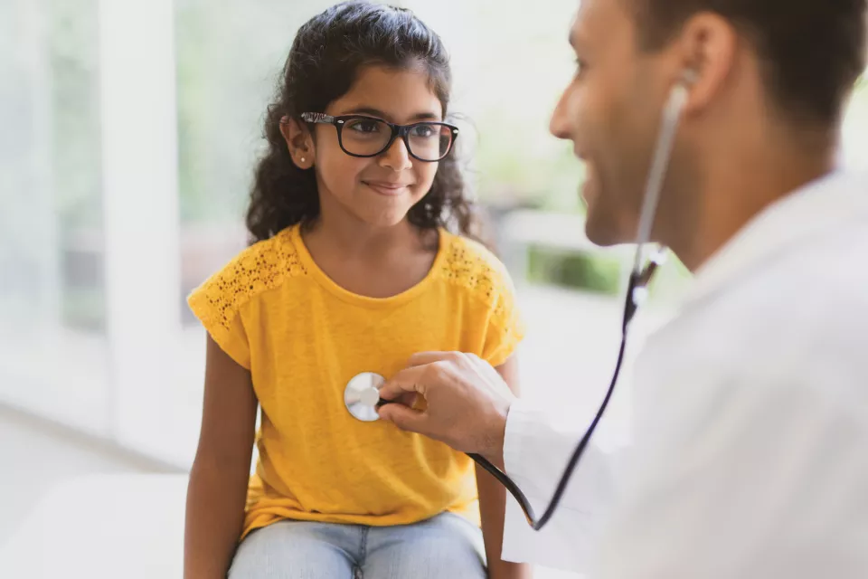 A little girl with medium-dark skin tone smiles as a doctor listens to her chest with a stethoscope
