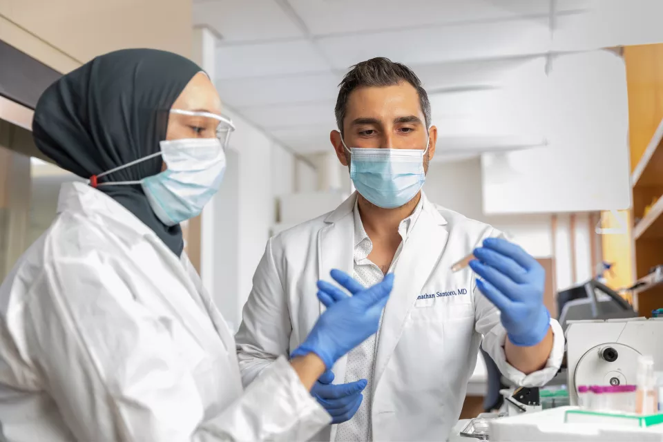 A woman with medium-light skin tone wearing a hijab shows a sample tube to a man with medium skin tone. Both wear lab coats and face masks.