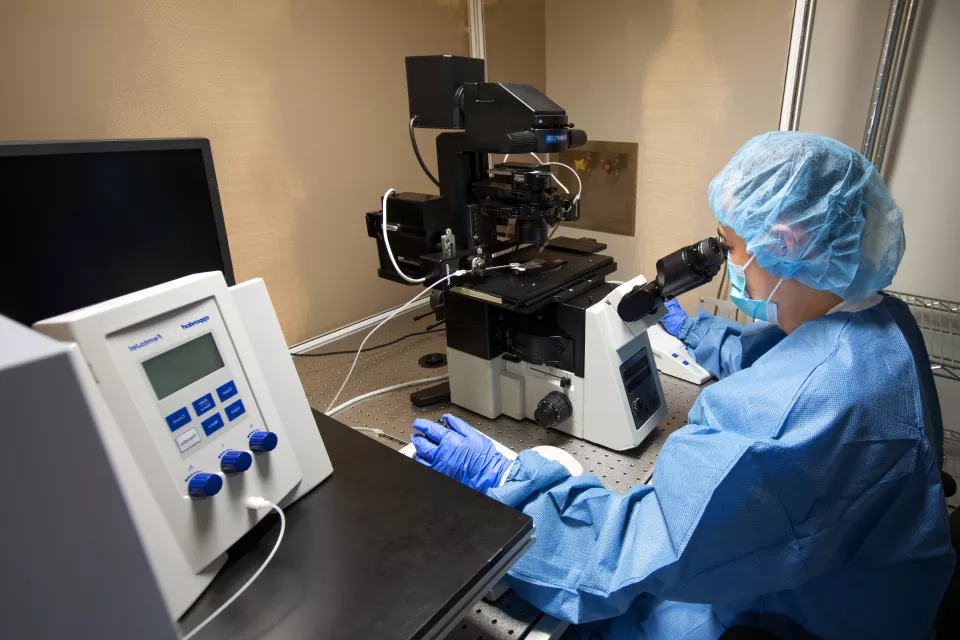 A woman wearing a medical mask and blue protective wear looks into the viewfinder of a large microscope connected to a device