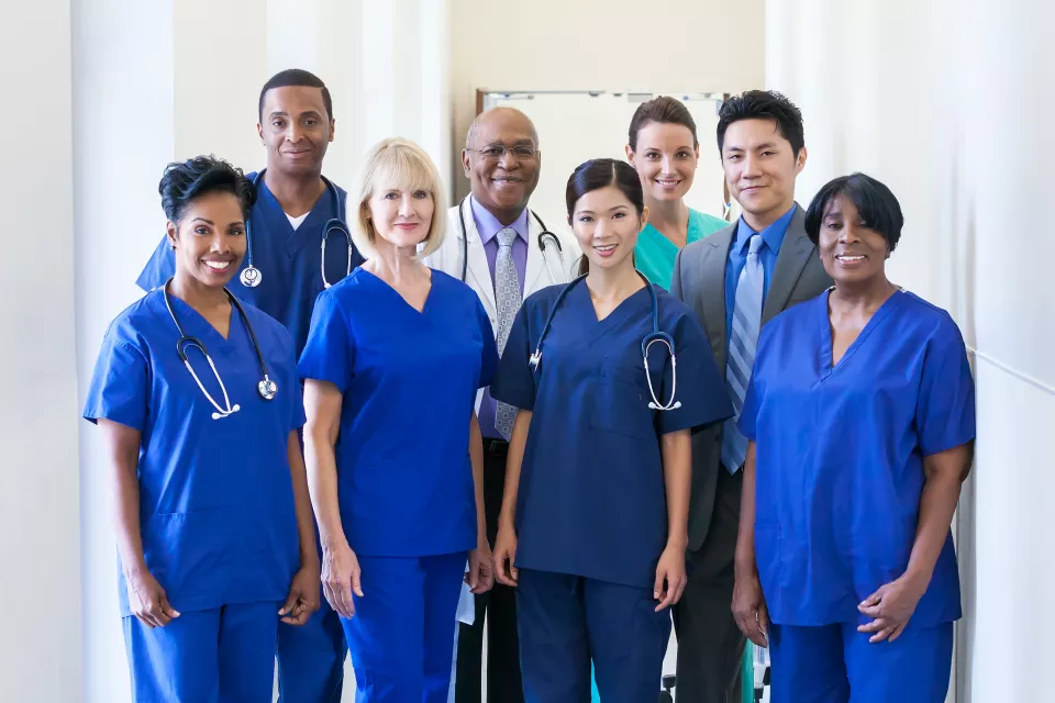 A group of 8 medical professionals of varying ethnicities smile. Six wear scrubs, one a suit, and one a lab coat and tie.