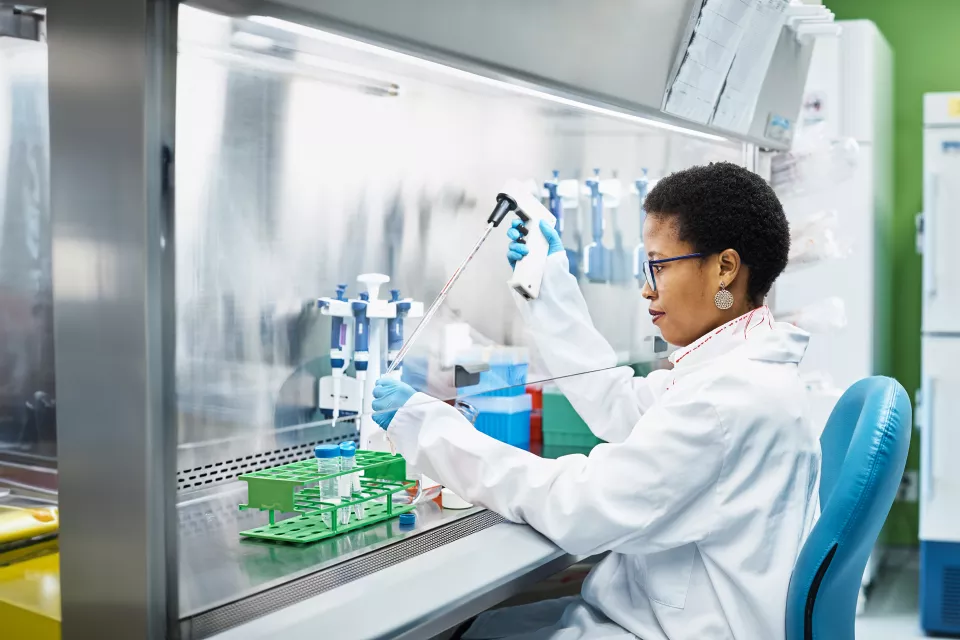 A woman with medium dark skin tone in a lab coat uses a pipetting device in a laboratory fume hood. 