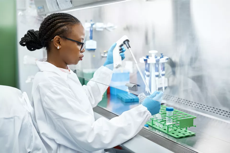 A woman with dark skin tone and braids tucked into a bun wears a lab coat and uses a pipette device in a laboratory fume hood. 