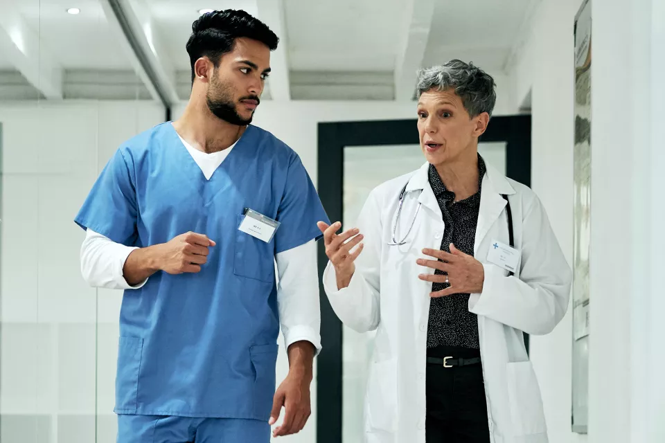 A medium-light skin toned woman with short gray hair wearing a lab coat and stethoscope talks to a medium skin toned man in blue scrubs as they walk. 