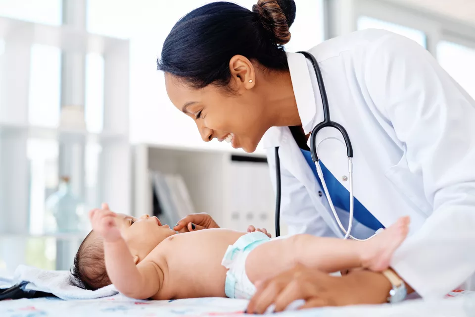 A medium skin toned woman in a lab coat and stethoscope smiles and examines a medium-light skin toned baby in a diaper. 