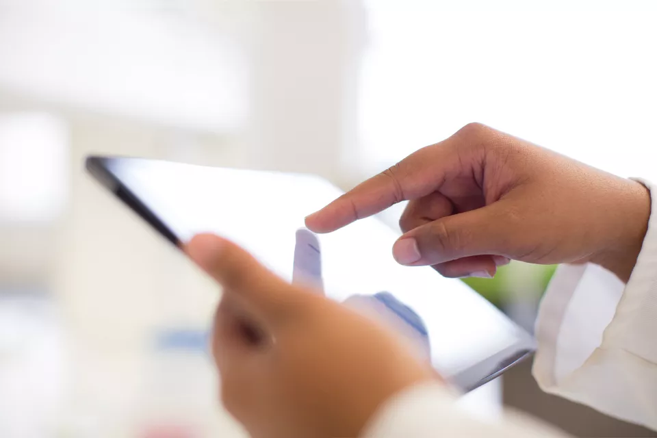 A person in a lab coat uses a digital tablet. Only medium skin toned hands are visible.