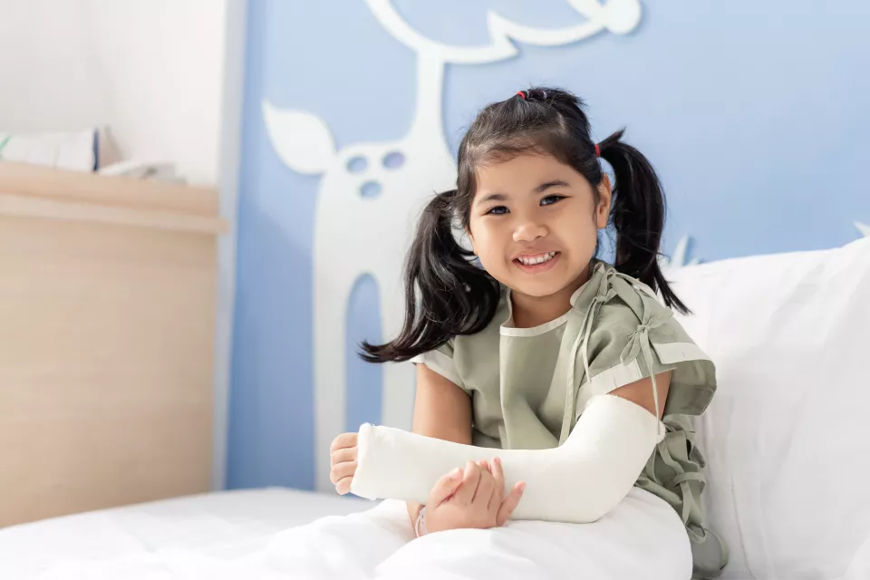 A girl with medium skin tone sits in a hospital bed, smiling and cradling her casted arm. She wears pigtails and a hospital gown.