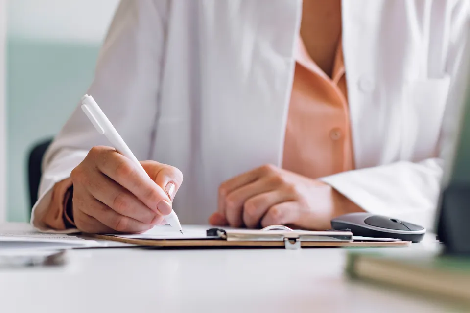 A view of the hands of a woman in a lab coat writing on a medical chart. 