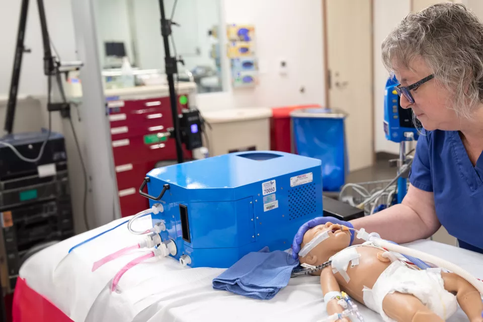 A light skin toned woman in blue scrubs leans over a simulated model of an infant laying on a hospital bed. 