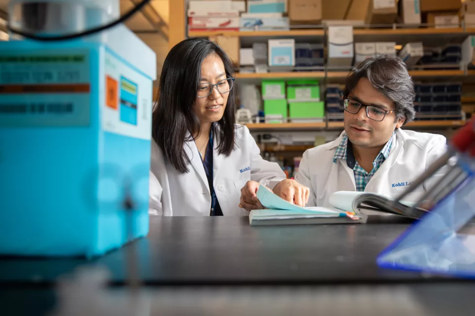 A medium skin toned woman points at a book that a medium skin toned man is looking at in a research laboratory. Both wear lab coats. 