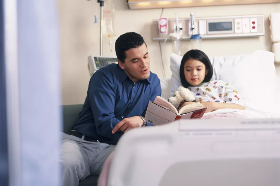 A medium-light skin toned man sits in a chair next to a hospital bed, reading a book to a medium-light skin toned girl holding a stuffed dog.