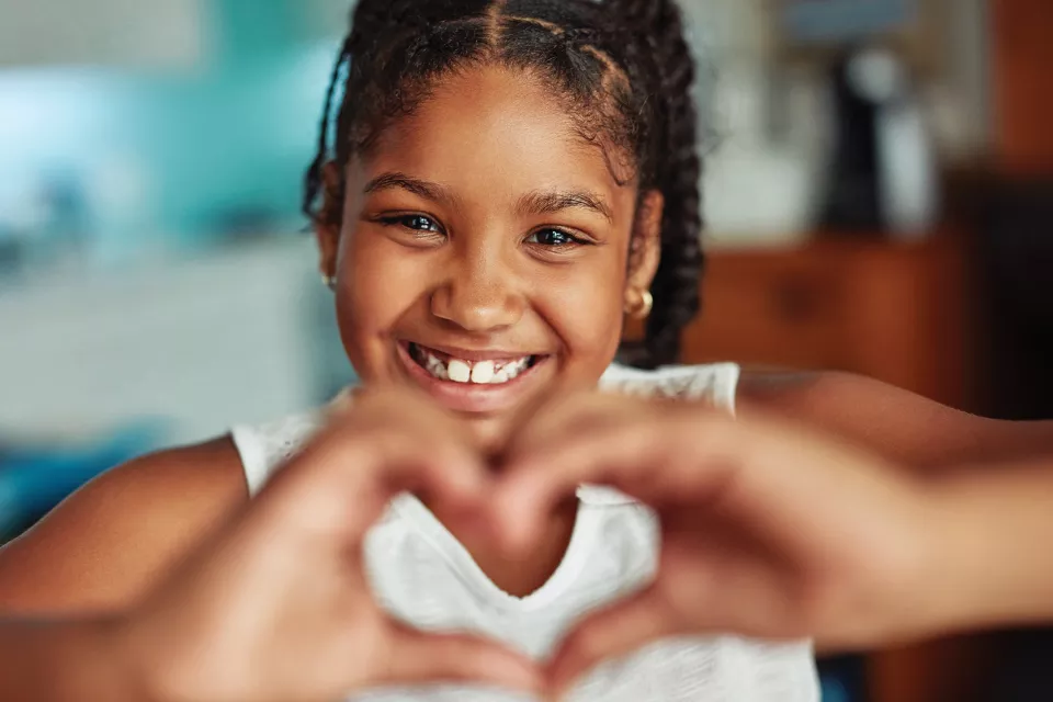 A girl with medium-dark skin tone smiles and makes a heart shape with her hands. 