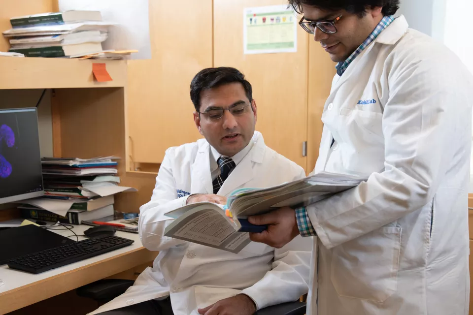 A man seated at a computer points at a book that a standing man is holding. Both have medium skin tone and wear lab coats.