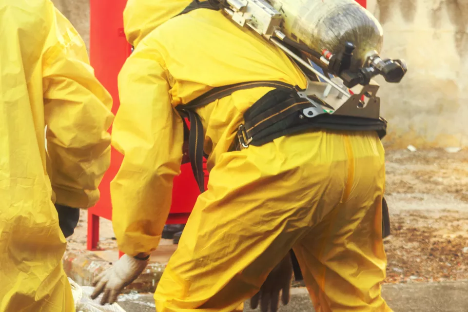 Three first responders wearing yellow HAZMAT suits and oxygen tanks bending down during drill