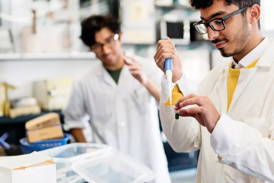 Student with medium skin tone and dark hair wearing black glasses and white lab coat learns how to use special laboratory equipment with professor watching in background