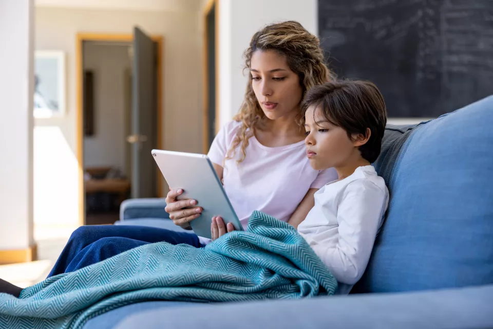 A medium skin-toned woman and a medium skin-toned child sit on a sofa looking at a tablet