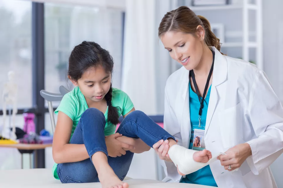 A light skin-toned woman in a lab coat and scrubs examines the bandaged ankle of a medium-light skin-toned girl seated on an exam table.