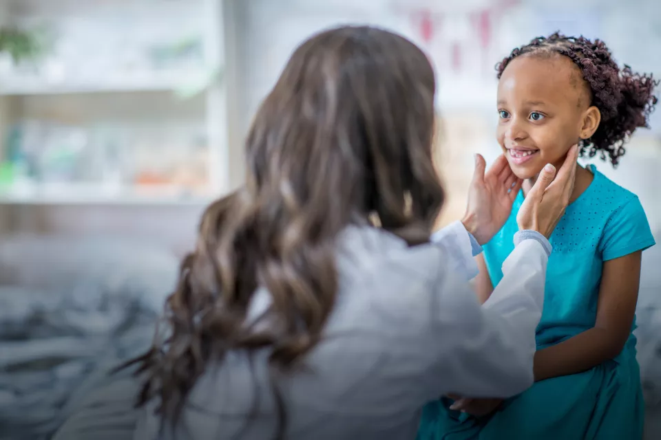 Young patient with medium skin tone and dark hair smiles as health care provider kneels in front of her and manually examines her neck