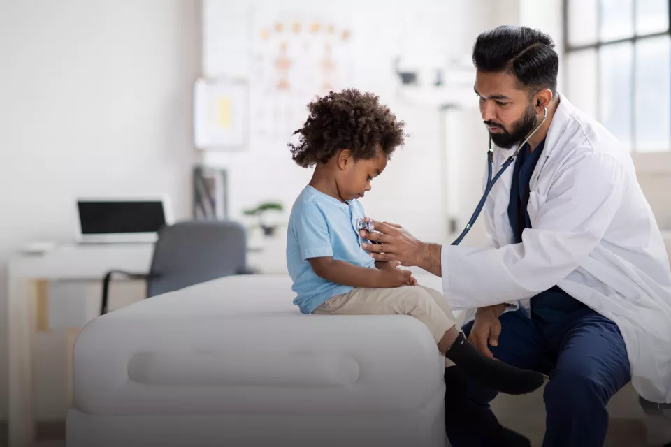 A male health care professional with medium skin tone and dark facial hair wearing a white lab coat uses a stethoscope to listen to the chest of a young patient with dark skin tone and curly brown hair in a medical office setting