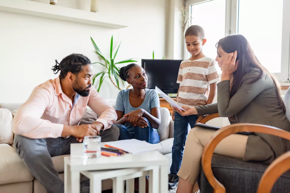 A man and woman sit on a couch, looking at a woman facing them in a chair, while a child stands between them.