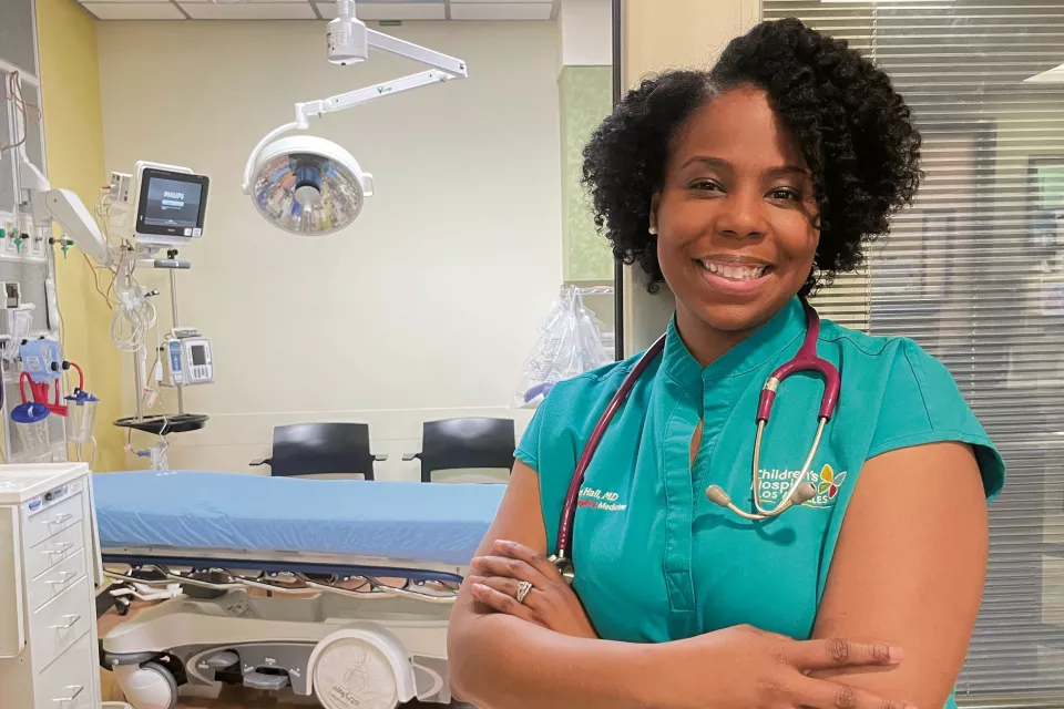 Female doctor with dark skin tone and dark hair smiles with arms crossed in hospital room