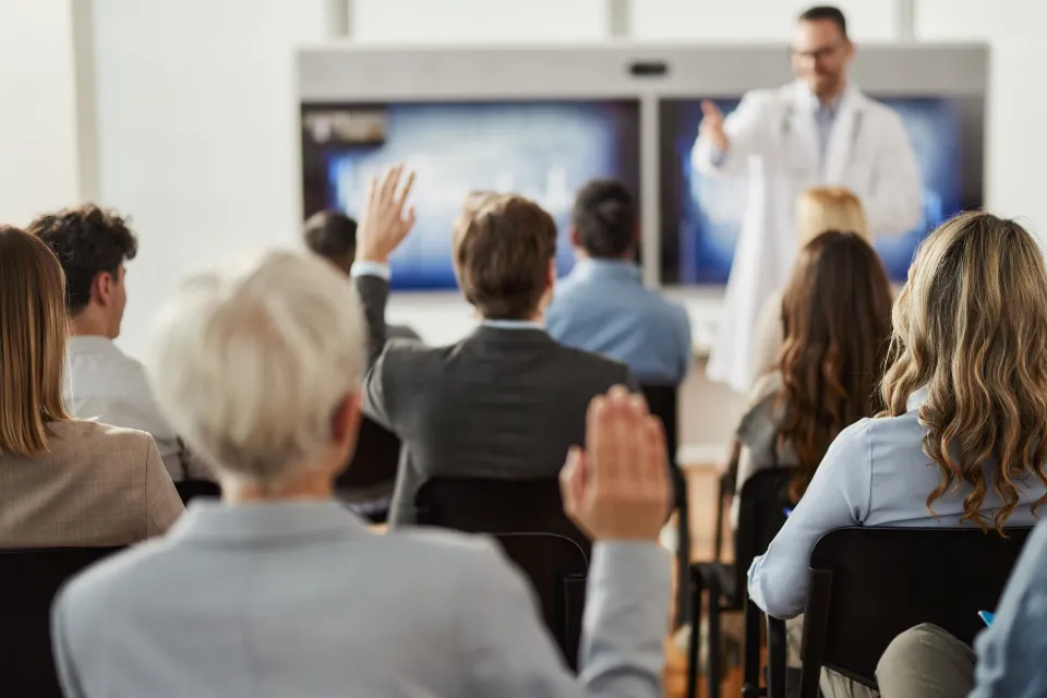 Man with light skin tone wearing lab coat and glasses instructs group of professionally dressed adult students in classroom setting