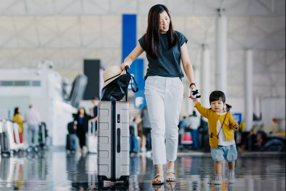 A mother with light skin tone and dark hair holds her young daughter's hand as they walk through an airport terminal