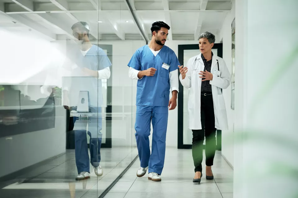 A medium-light skin toned woman with short gray hair wearing a lab coat and stethoscope talks to a medium skin toned man in blue scrubs as they walk. 