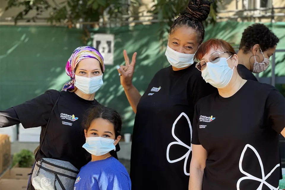 3 women in black CHLA T-shirts and a child in a blue shirt, all wearing surgical masks, smile at the camera. The medium-light skin-toned woman wears a colorful head scarf and the medium-dark skin-toned woman makes a piece sign with her fingers.  