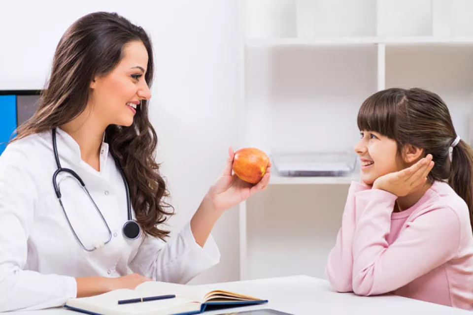 A medium-light skin-toned woman in a lab coat with a stethoscope smiles and holds an apple up to a medium-light skin-toned smiling girl.