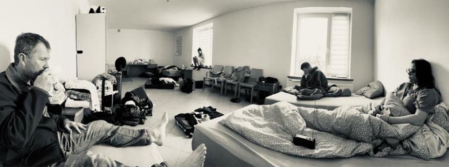 In black and white, 4 tired-looking people sit around a room containing beds, chairs and luggage. They are resting, eating or reading. 