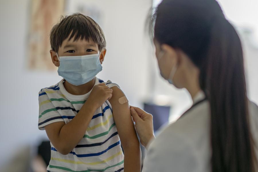 A young medium skin-toned boy wearing a surgical mask holds up his sleeve as a woman in a lab coat puts a bandage on his upper arm