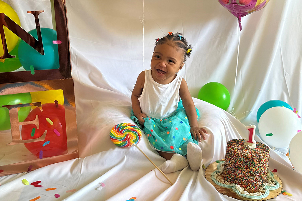 Ciara, a medium skin-toned toddler, smiles as she sits next to a rainbow swirl lollipop and a birthday cake topped with a number one candle