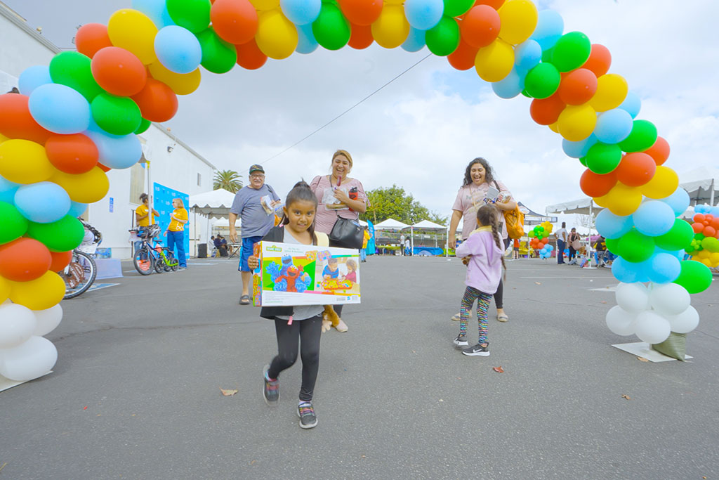 Young girl holding Sesame Street toy walks beneath an arch of rainbow balloons 