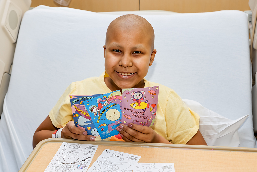 Young patient smiles in their hospital bed as they hold Valentine's Day cards 