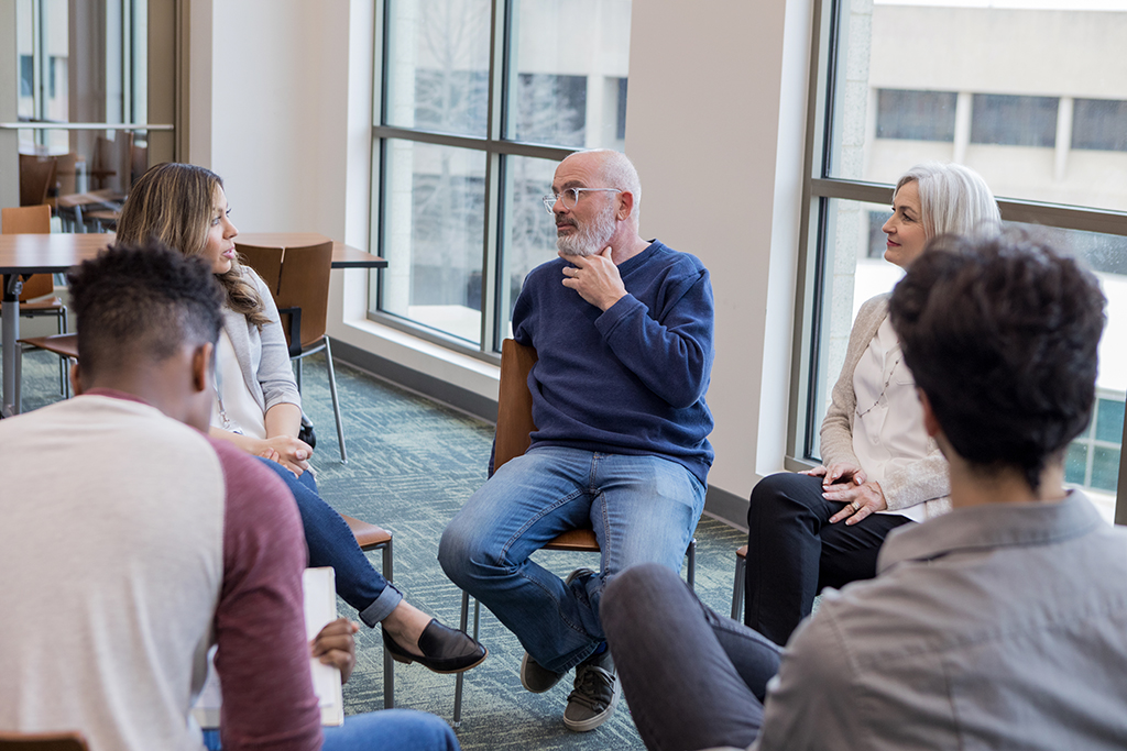 Group of five casually dressed adults engage in roundtable discussion in hospital conference room setting